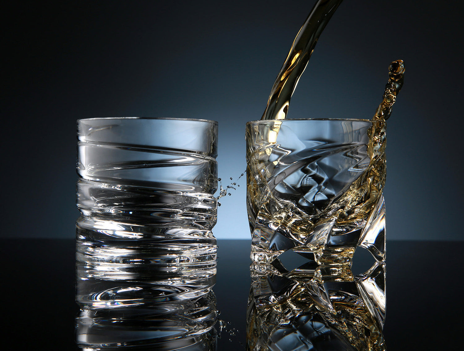 Two crystal shot glasses with spiral designs, one being filled with amber liquid, causing a splash, placed on a reflective surface against a dark background.