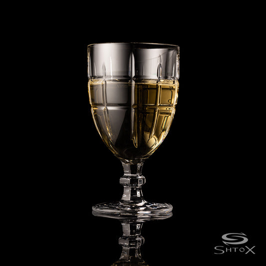 Crystal rotating wine glass with a bold geometric pattern, filled with light golden liquid, set against a black background with the Shtox logo at the bottom.
