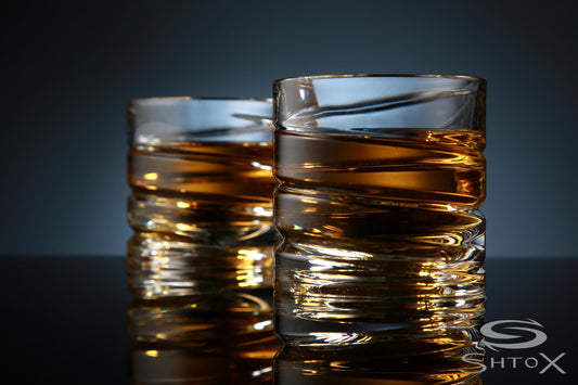 Two crystal rotating shot glasses with a spiral design, filled with amber liquid, placed on a reflective surface against a dark background, featuring the Shtox logo in the corner.