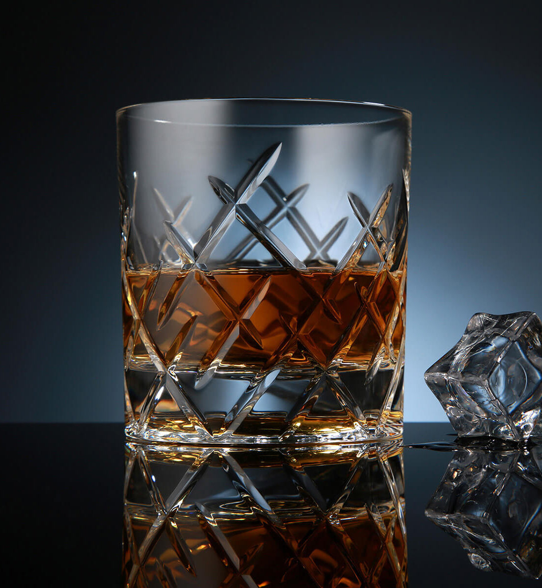 Crystal whiskey rotating glass with a diamond-cut pattern, filled with amber whiskey, placed on a reflective surface next to a faceted ice cube, set against a dark blue background.