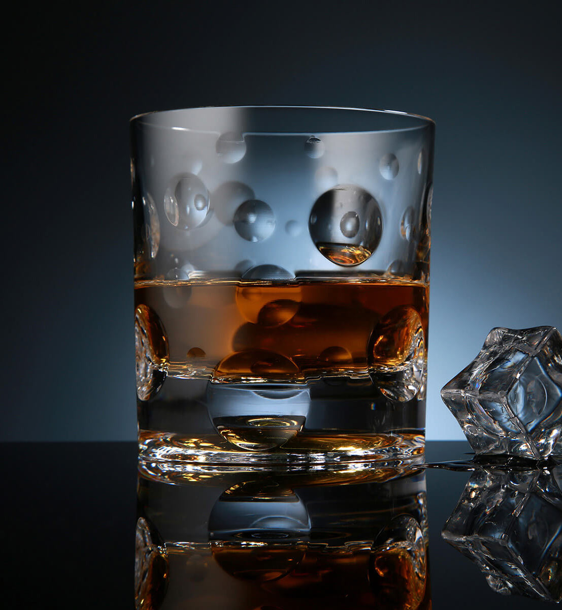 Crystal whiskey rotating glass with an embossed bubble pattern, filled with amber whiskey, placed on a reflective surface next to a faceted ice cube, set against a dark blue background.