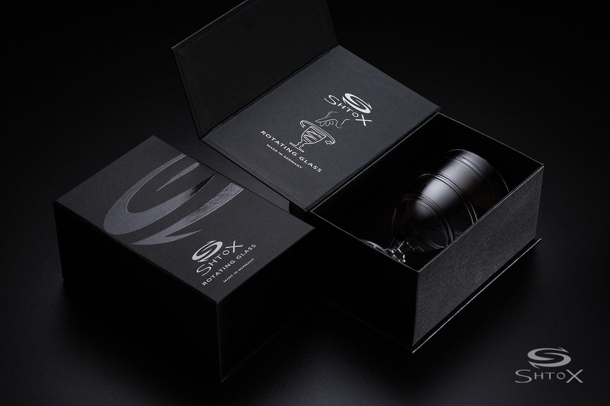 Luxury packaging of a Shtox rotating wine glass, featuring sleek black boxes with silver branding and a partially visible glass inside, set against a dark background.