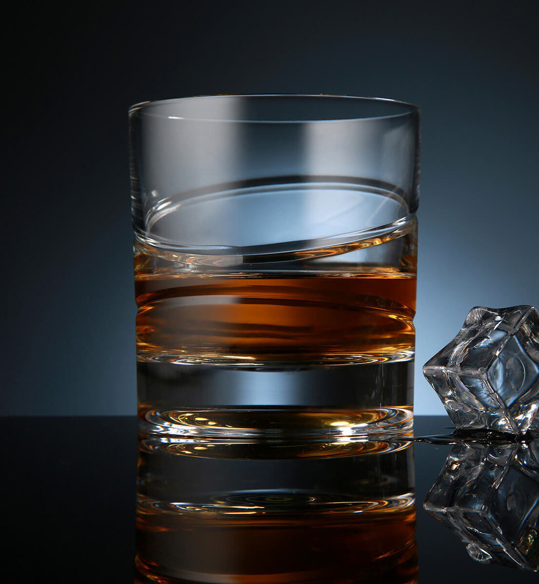 Crystal whiskey rotating glass with a sleek, minimalist design, filled with amber whiskey, placed on a reflective surface next to a faceted ice cube, set against a dark blue background.