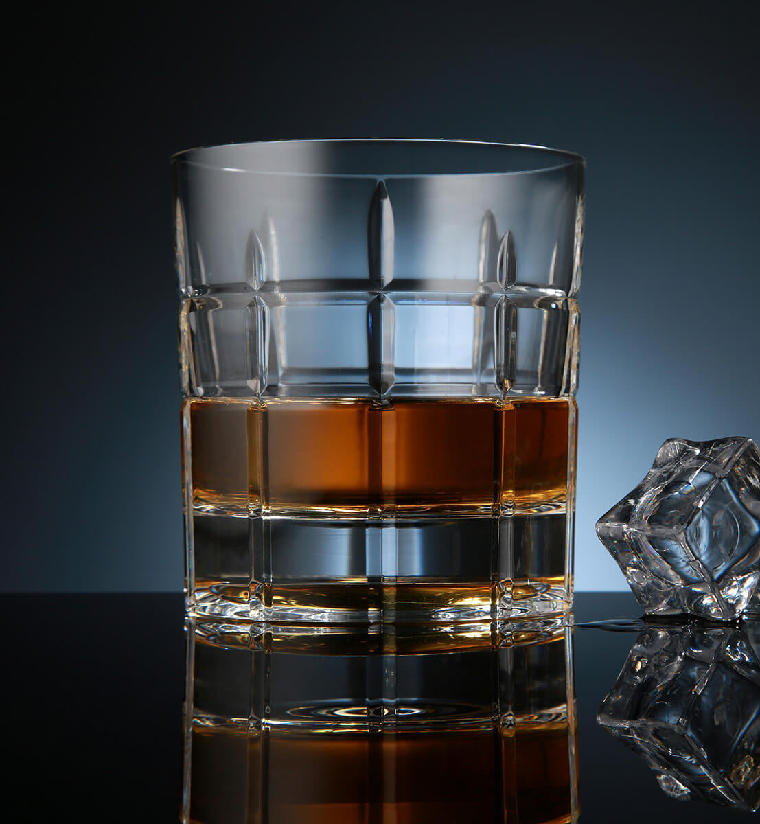 Crystal whiskey rotating glass with a square-cut grid pattern, filled with amber whiskey, placed on a reflective surface next to a faceted ice cube, set against a dark blue background.
