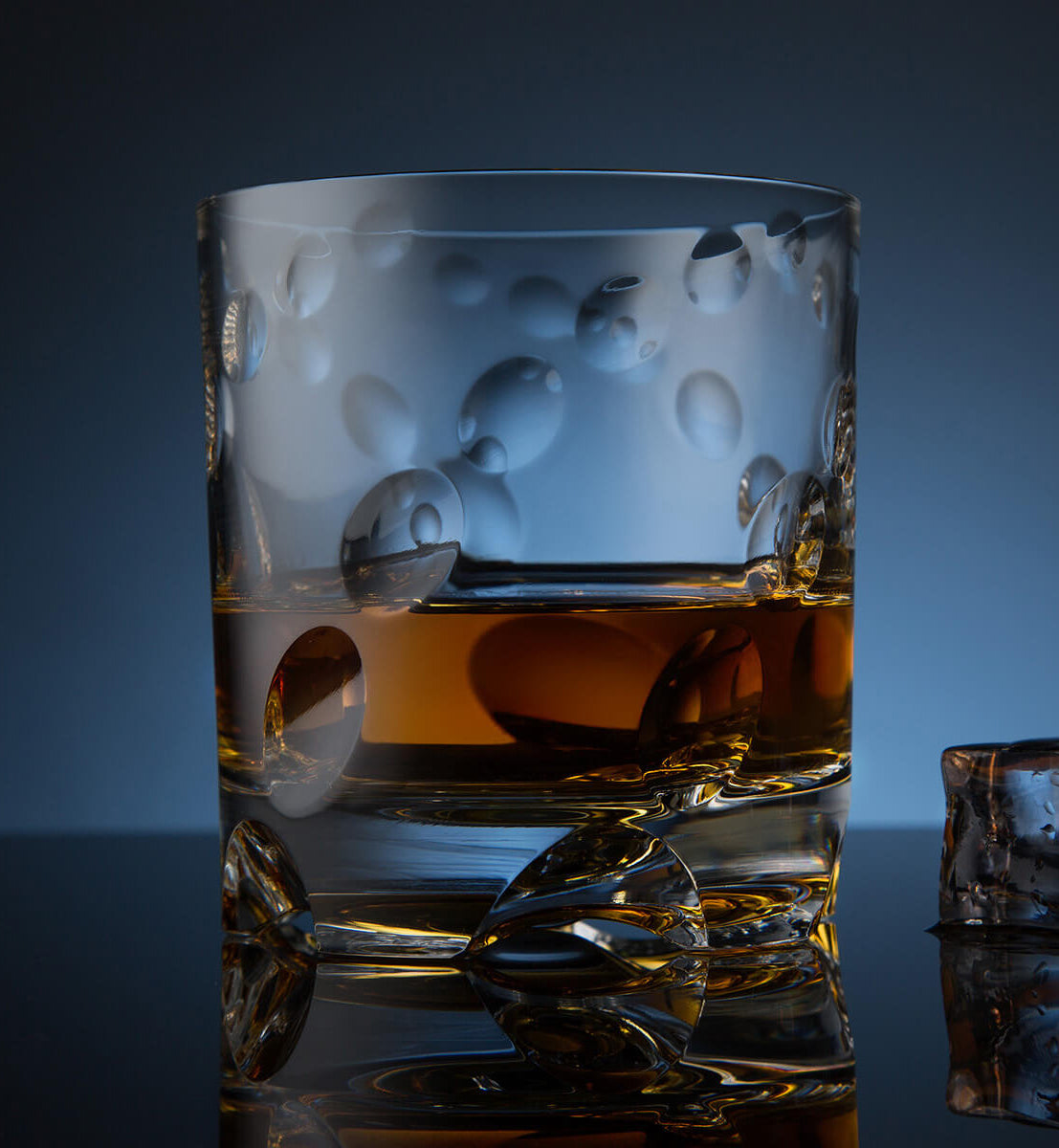 Crystal whiskey rotating glass with an embossed bubble pattern, filled with whiskey, placed on a reflective surface next to a faceted ice cube, set against a dark blue background.