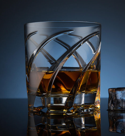 Crystal whiskey rotating glass with a dynamic curved etched design, filled with amber whiskey, placed on a reflective surface next to a faceted ice cube, set against a dark blue background.