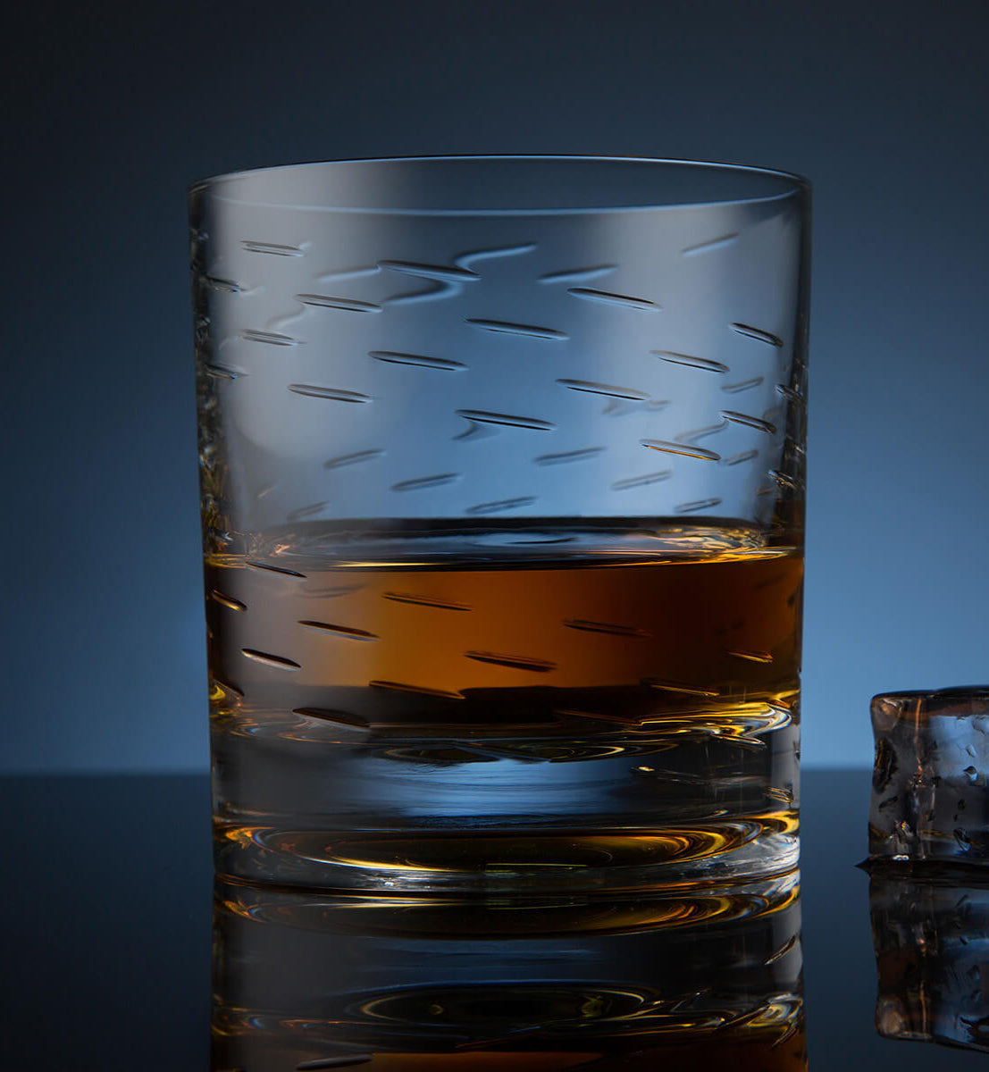 Crystal whiskey rotating glass with etched diagonal lines, filled with amber whiskey, placed on a reflective surface next to a faceted ice cube, set against a dark blue background.
