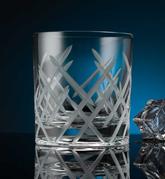 Crystal whiskey rotating glass with intricate frosted geometric pattern, placed on a reflective surface next to a faceted ice cube, set against a dark blue background.