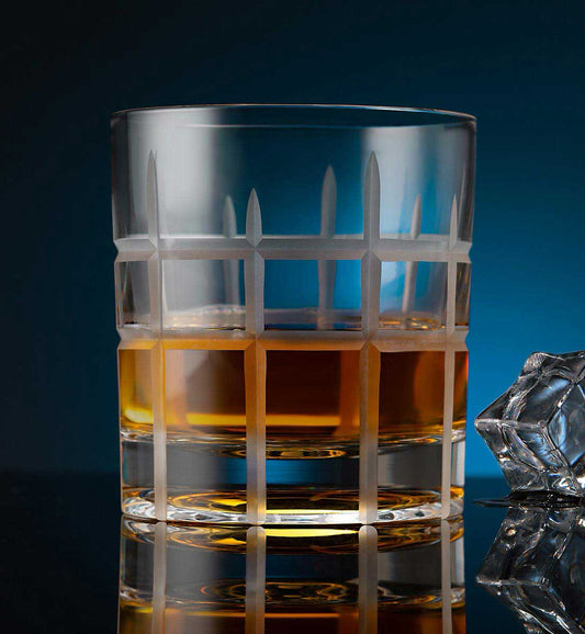 Crystal whiskey rotating glass with a matte geometric grid pattern, filled with amber liquid, placed on a reflective surface next to a faceted ice cube, set against a dark blue background.