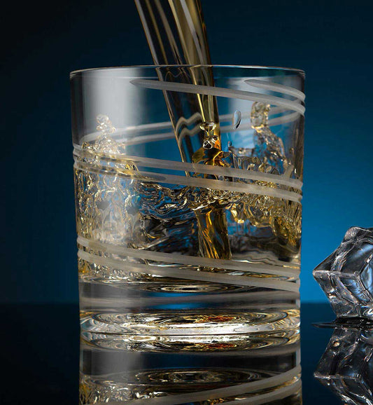 Crystal whiskey rotating glass with frosted diagonal lines, being filled with whiskey, placed on a reflective surface next to a faceted ice cube, set against a dark blue background.