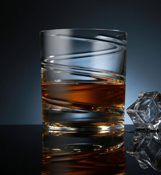 Crystal whiskey rotating glass with a spiral etched design, filled with amber whiskey, placed on a reflective surface next to a faceted ice cube, set against a dark blue background.