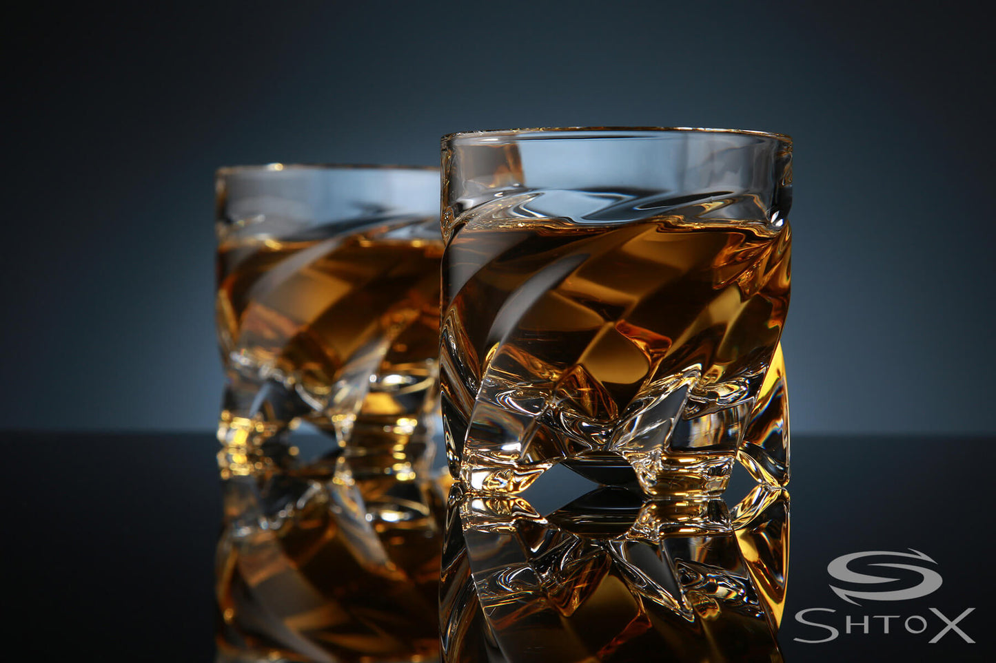 Two crystal rotating shot glasses with a twisted design, filled with amber liquid, placed on a reflective surface against a dark background, featuring the Shtox logo in the corner.