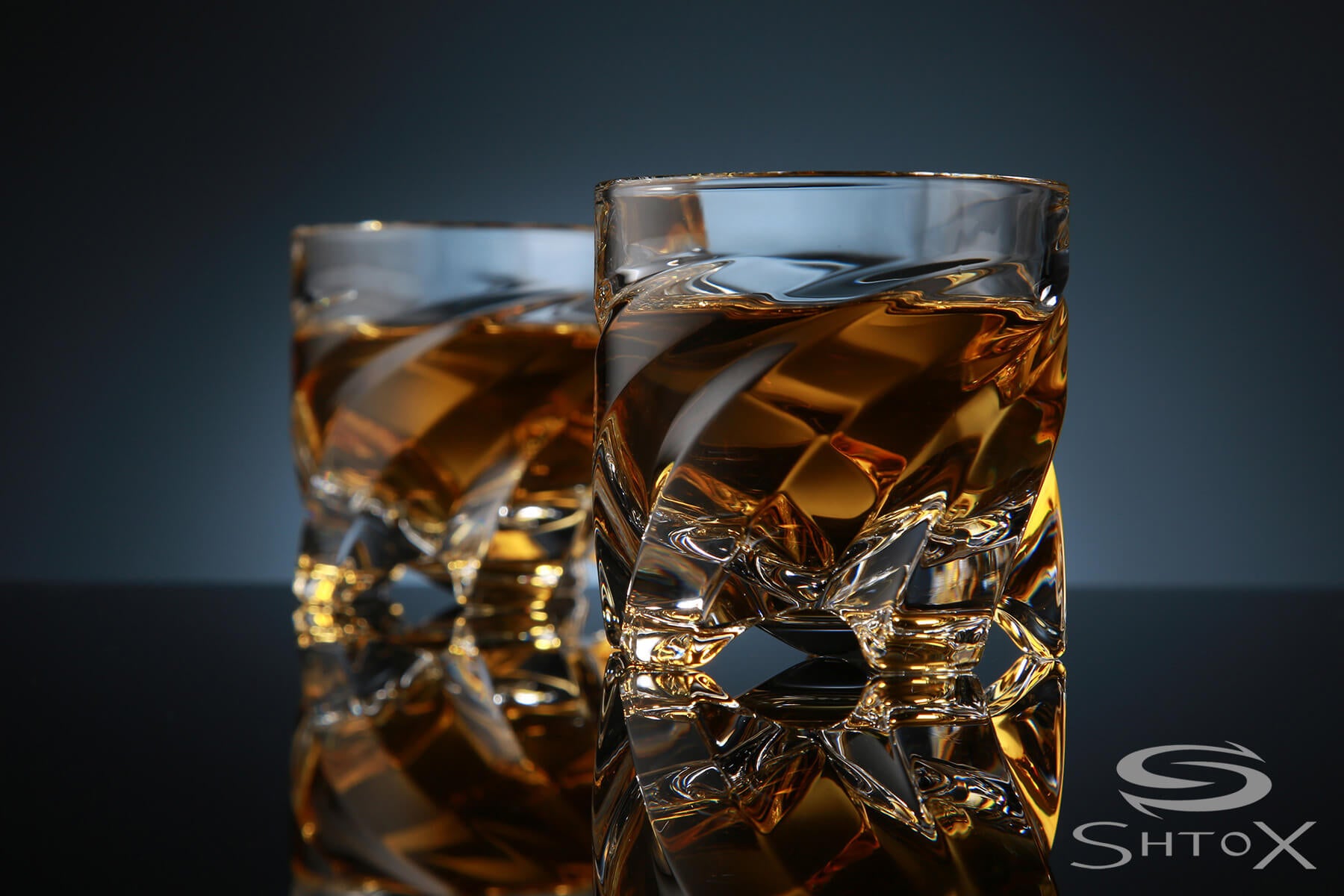 Two crystal rotating shot glasses with a twisted design, filled with amber liquid, placed on a reflective surface against a dark background, featuring the Shtox logo in the corner.