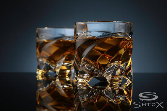 Two crystal rotating shot glasses with a twisted design, filled with amber liquid, placed on a reflective surface against a dark background, featuring the Shtox logo in the corner.