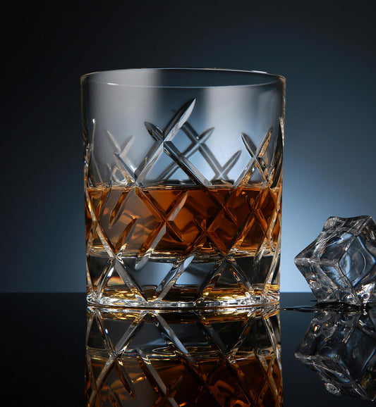 Crystal whiskey rotating glass with a diamond-cut pattern, filled with amber whiskey, placed on a reflective surface next to a faceted ice cube, set against a dark blue background.