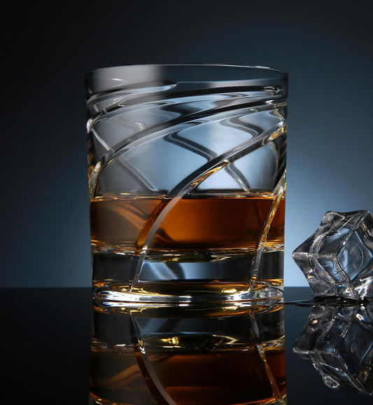 Crystal whiskey rotating glass with a curved spiral etched design, filled with amber whiskey, placed on a reflective surface next to a faceted ice cube, set against a dark blue background.