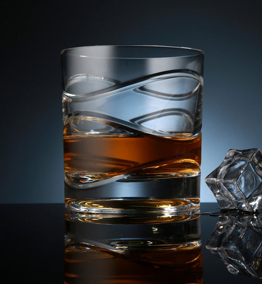 Crystal whiskey rotating glass with a wave-like etched design, filled with amber whiskey, placed on a reflective surface next to a faceted ice cube, set against a dark blue background.