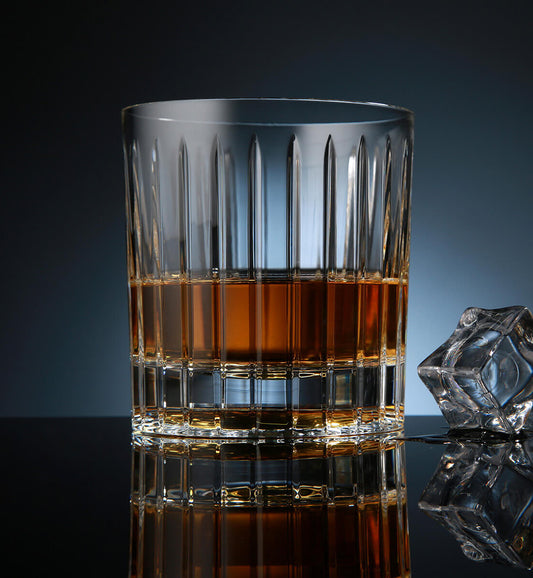 Crystal whiskey rotating glass with vertical cut grooves, filled with amber whiskey, placed on a reflective surface next to a faceted ice cube, set against a dark blue background.