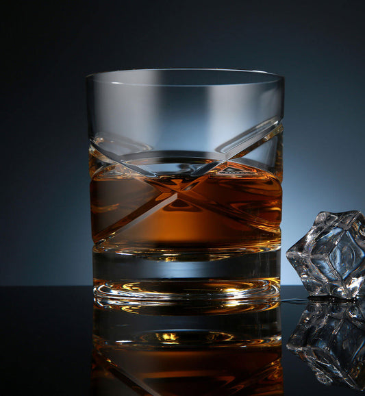 Crystal whiskey rotating glass with amber liquid, featuring a modern geometric design and a reflective surface, alongside a faceted ice cube on a dark background.