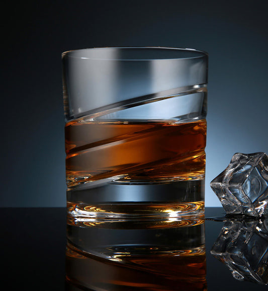 Crystal whiskey rotating glass with a smooth spiral etched design, filled with amber whiskey, placed on a reflective surface next to a faceted ice cube, set against a dark blue background.