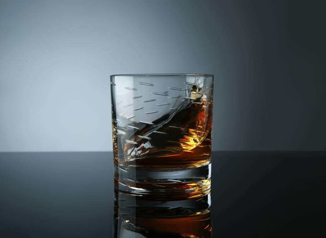 Crystal whiskey tumbler with etched diagonal lines, filled with amber liquid, placed on a reflective surface against a soft gradient background.
